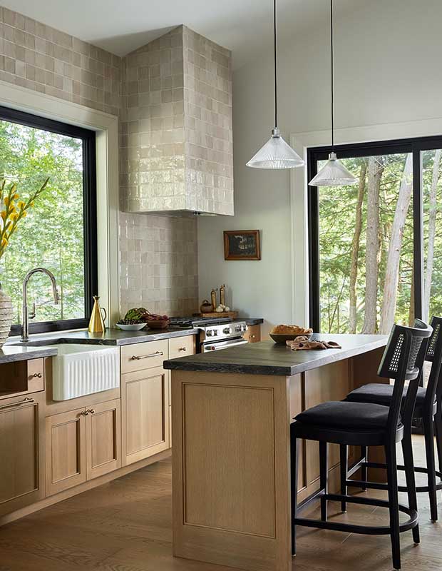 A kitchen with light-wood elements, glassy beige tile, a small island, and large windows that look out into the forest. serene muskoka bunkie