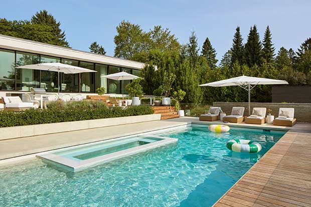 A bright blue pool with a built-in hot tub and large umbrella hanging over deck chairs. unique backyard pools
