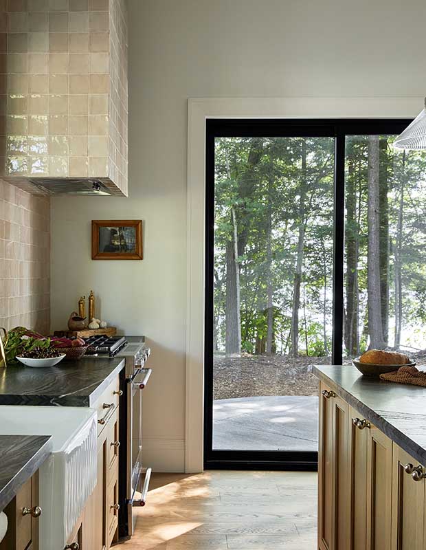 A kitchen with light-wood floors and glassy beige tile looks out into a forest. serene muskoka bunkie