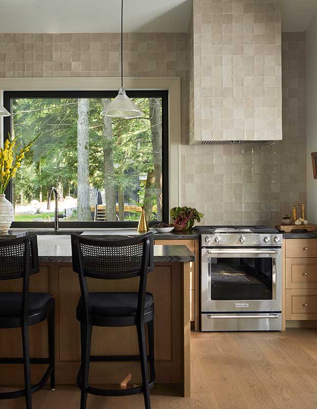 A kitchen with glassy beige tile walls, light wood flooring and island, black hardware, and a large window that looks out into the forest. serene muskoka bunkie