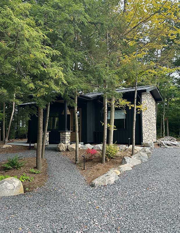 A black-walled bunkie with stone siding stands in the trees with rock paths surrounding it. serene muskoka bunkie