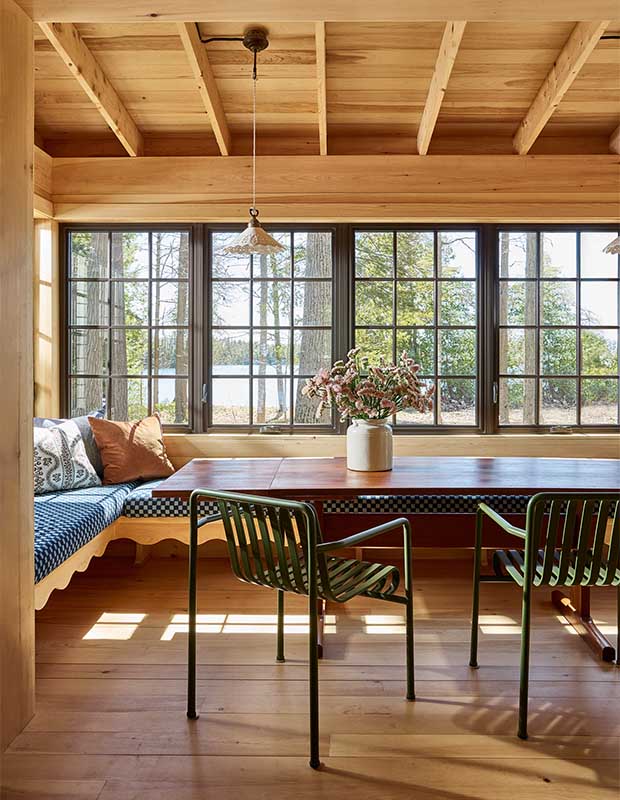 A pine dining nook with banquet seating and a long dark-wood table. Blue checked cushions and deep green wire chairs are accents.wood-clad family cottage