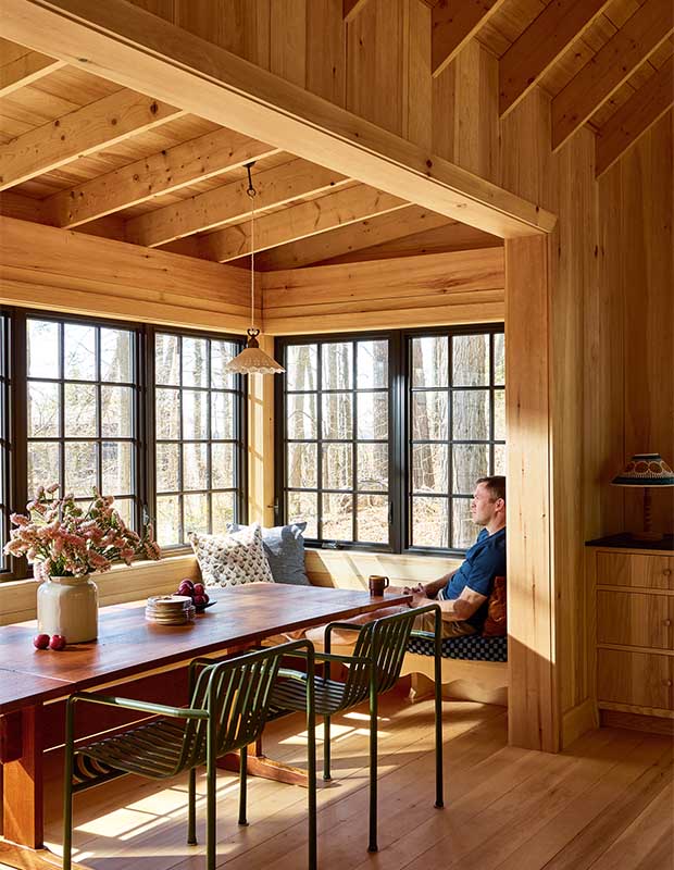 A caucasian man sits at a banquet seat in a dining nook, looking out the window with a cup of coffee. wood-clad family cottage