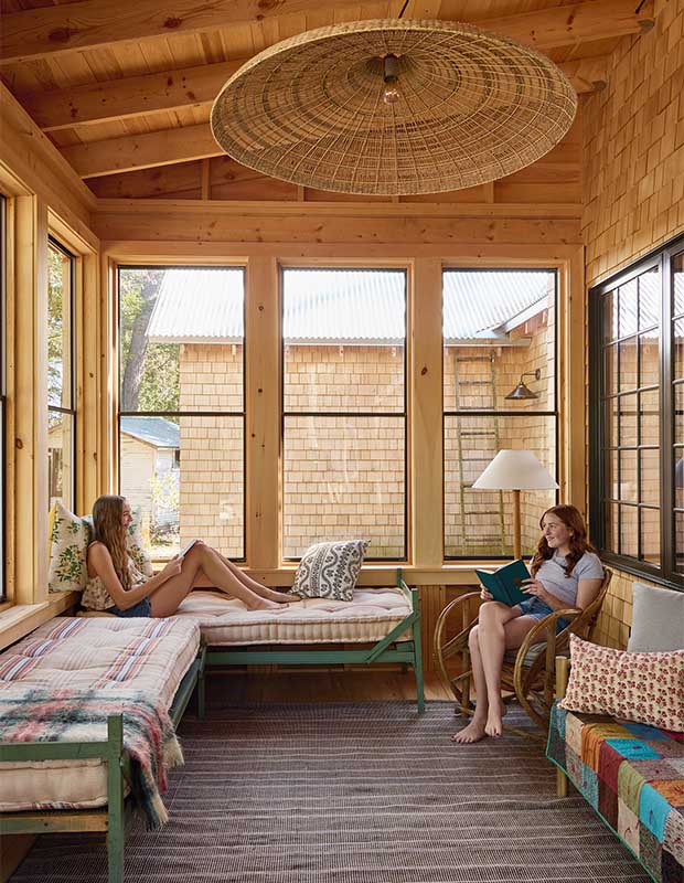 Two tween girls sit in a pine sunroom with benches with cushions and ornate wicker chairs. wood-clad family cottage