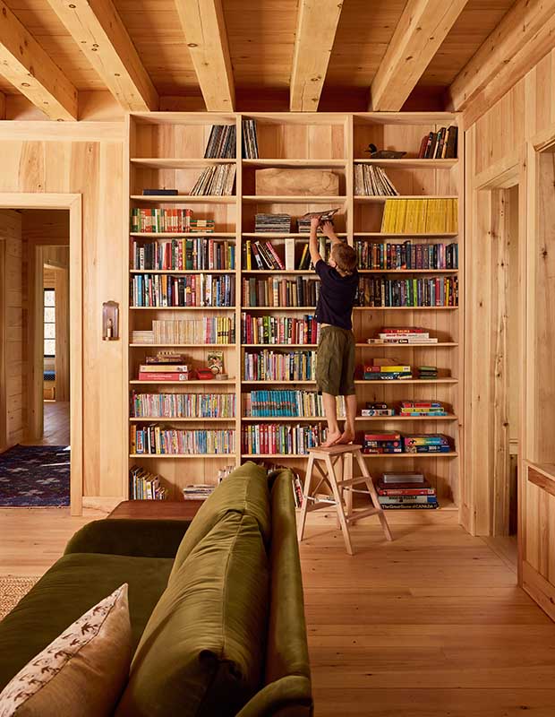 A child stands on a footstool in front of a floor-to-ceiling book case. wood-clad family cottage