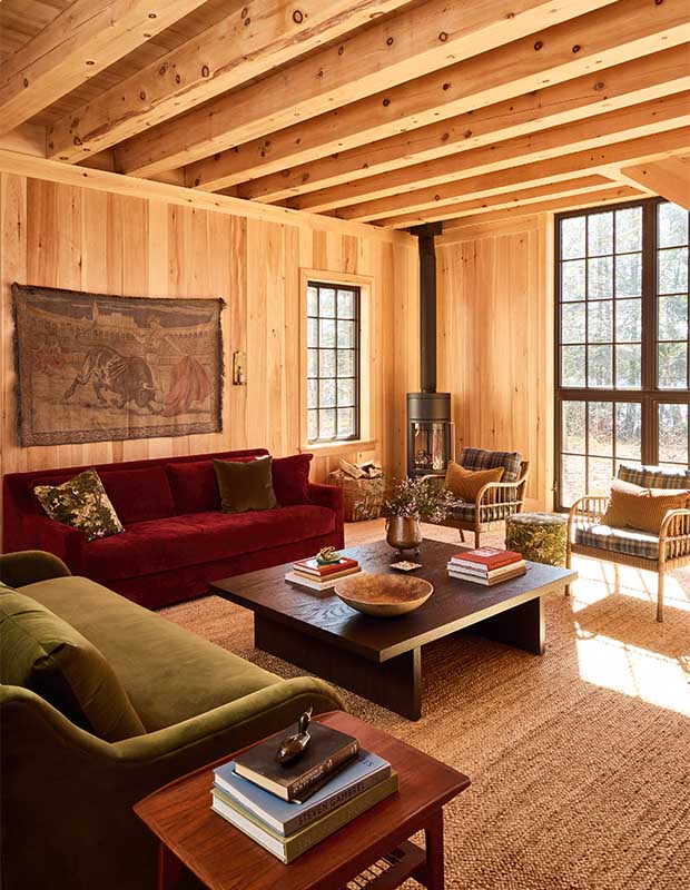 A pine lined living room with red and green couches, retro chairs, a woven wall hanging, and airy light panelled windows. wood-clad family cottage