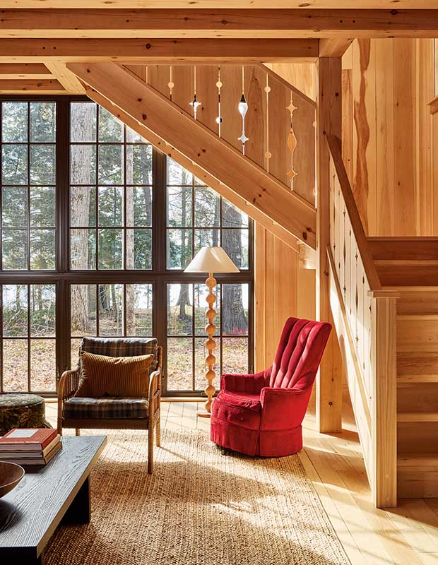 In a pine living room is a pained glass window. Pine stairs lead to an upper floor, accented with carved space. A red chair, as well as an old-school sitting chair and large rug sit on the main floor. wood-clad family cottage