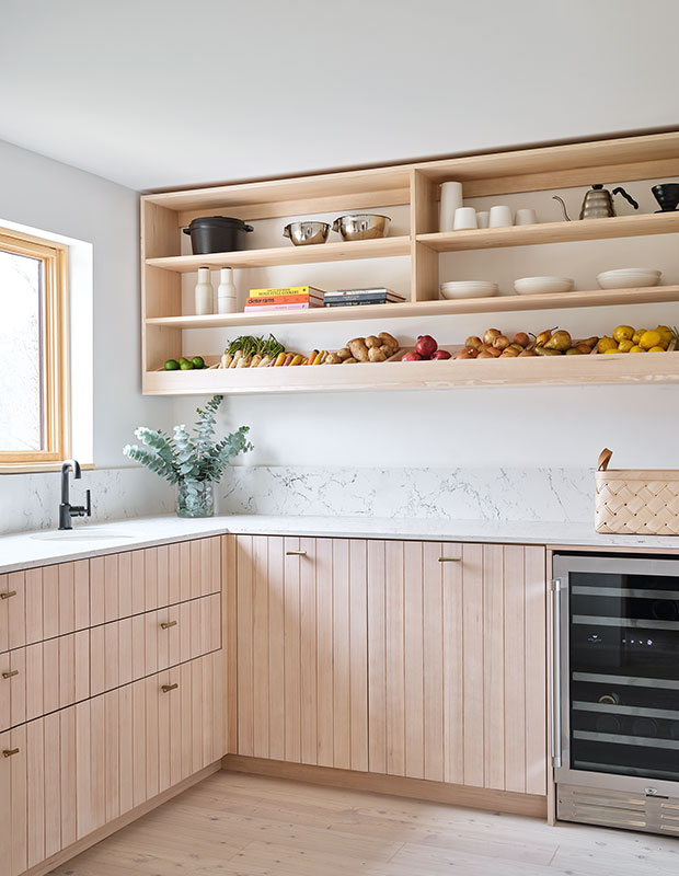 An image of the corner of a pantry in a Scandi-style farmhouse with blond wood cupboards and open shelves with cups, bowls, and vegetables.