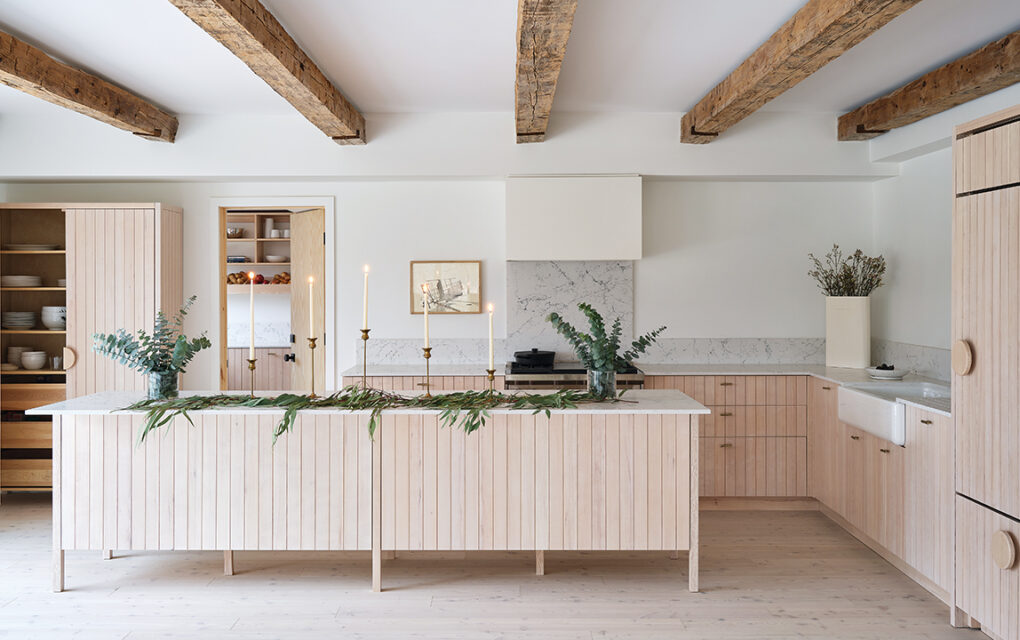 A photo of a kitchen in a Scandi-style farmhouse with a large island and cupboards in blond wood, exposed wooden beams and white walls.