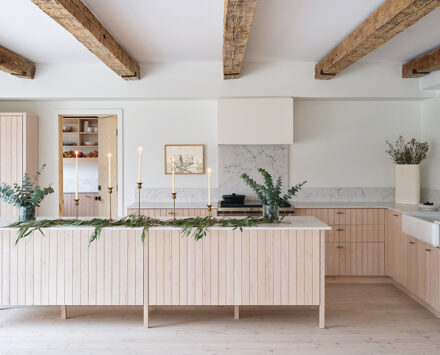 A photo of a kitchen in a Scandi-style farmhouse with a large island and cupboards in blond wood, exposed wooden beams and white walls.