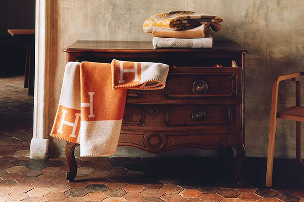 A stylish throw with a letter H motif in orange and white is flowing out of a wooden armoire.