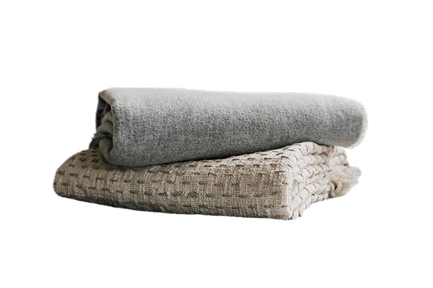 A folded stylish throw in beige with a folded grey throw stacked on top of it against a white background.