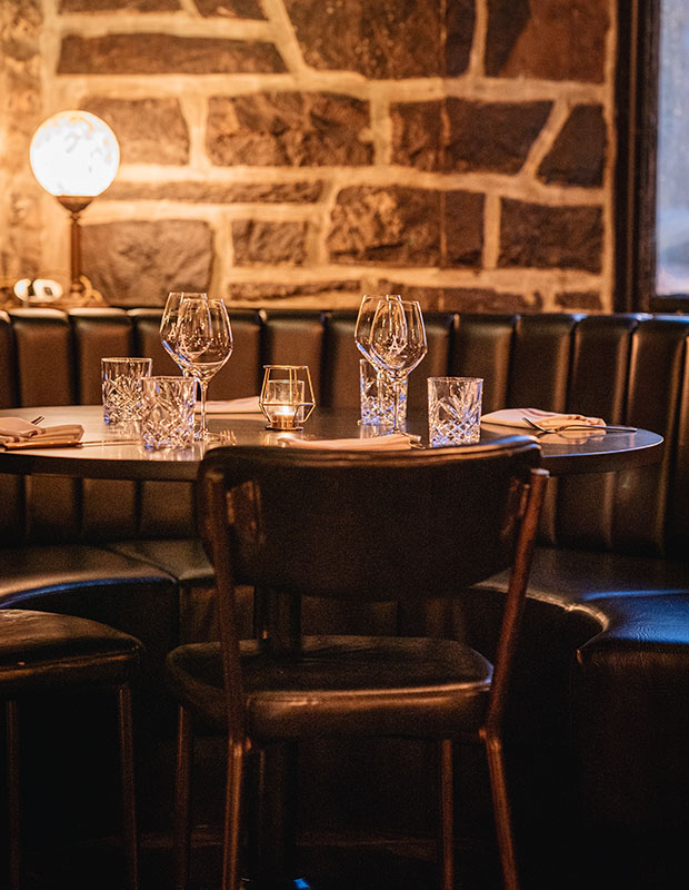 A photo of a table at Auberge Saint-Gabriel. A round light illuminates a dimly lit corner booth at a restaurant topped with clear wine glasses.