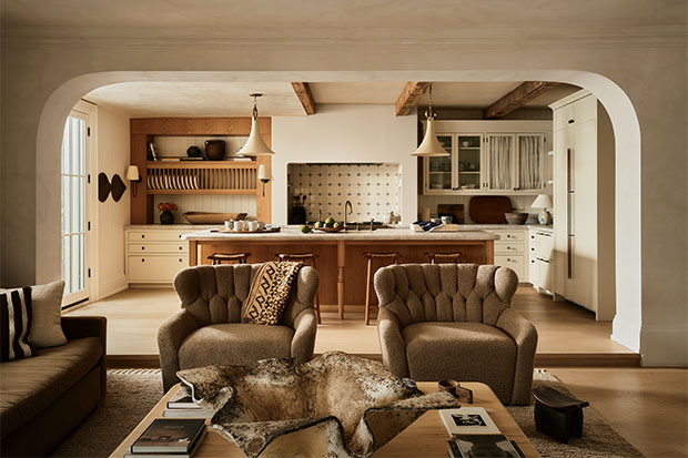 A photograph of a kitchen viewed from the living room. Two armchairs face the camera and a wood kitchen island sits behind them. Behind that is a range with cabinets on either side of it. The kitchen walls are painted white and there are large wooden beams crossing the ceiling.
