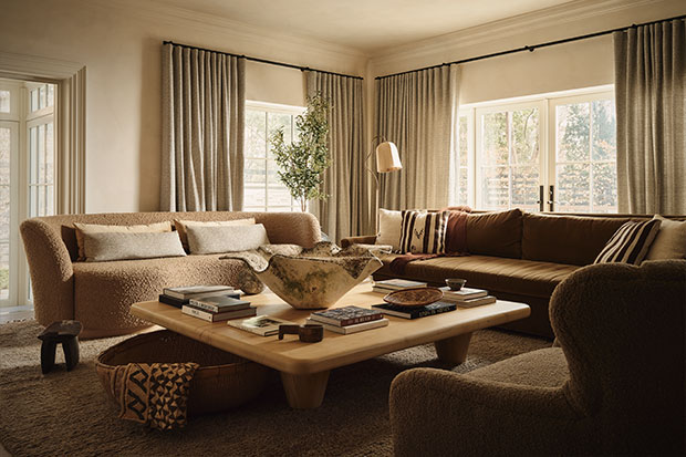A photograph of a living room. Two large windows and their curtains fill the walls. In the corner, a floor lamp stands between two couches. Both couches face a wooden table with a large decorative bowl in the middle of it.