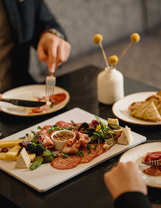 A photo of food at the AC Old Montreal hotel. A rectangular plate has an assortment of cheeses and cured meats and a small dish of mustard. It is set on a black table, and the hands of a guest are visible cutting a piece of meat on their plate with a knife and fork.