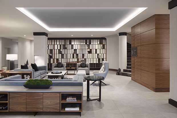 A photograph of the lobby of the AC Old Montreal hotel. A low counter with wooden cupboards and a grey countertop is in the foreground and a stylized shelving unit with rectangle motifs is in the background. The walls are white, and a seating area with several grey chairs and a grey couch occupies the centre of the room.