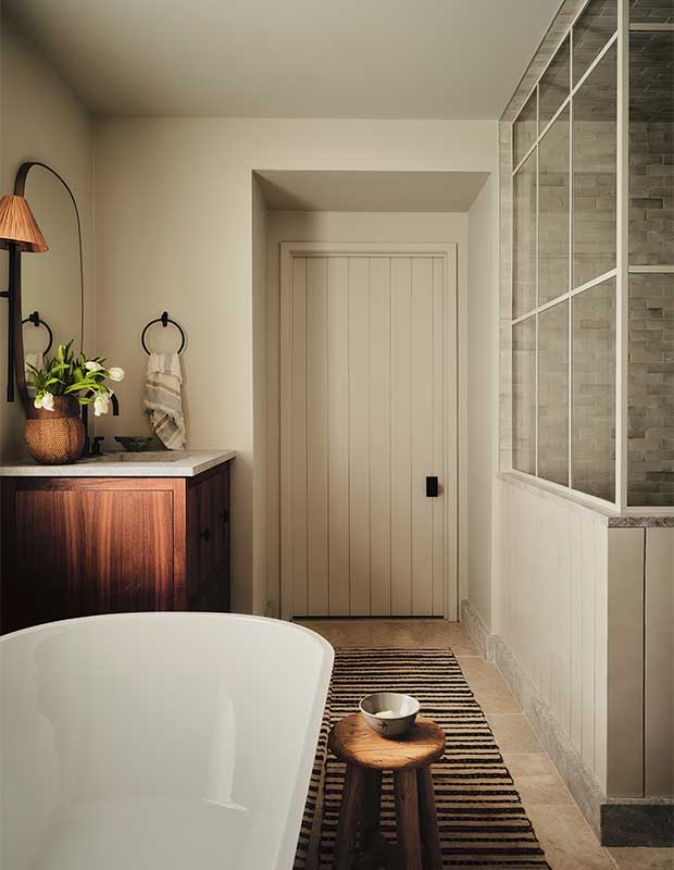 A photograph of the ensuite. On the left, a corner of a freestanding bathtub is visible, and a wooden vanity is attached to the wall. On the right, a glass-enclosed shower is visible. In the centre is a white door.