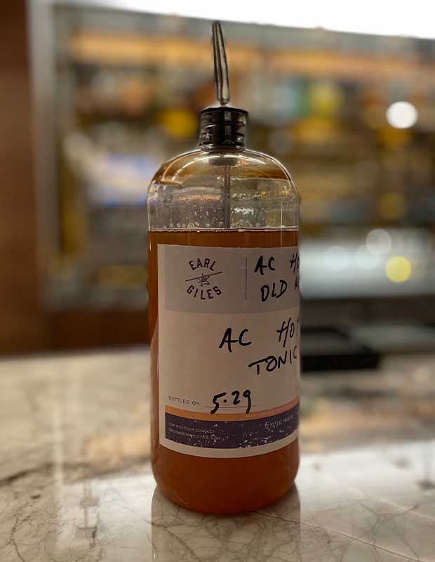 A bottle of tonic concentrate at the AC Old Montreal hotel. It is a clear bottle with a white label with ingredients written on it with a black marker and a pour spout attached. It is three quarters full of an orange liquid.