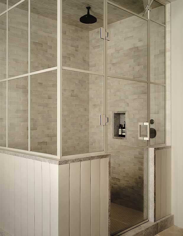 A photograph of the shower in the ensuite. It is enclosed with white wainscotting below glass panes framed by white muntins. The inside is covered in grey, rectangular tiles and there is a small niche with soap bottle in it.