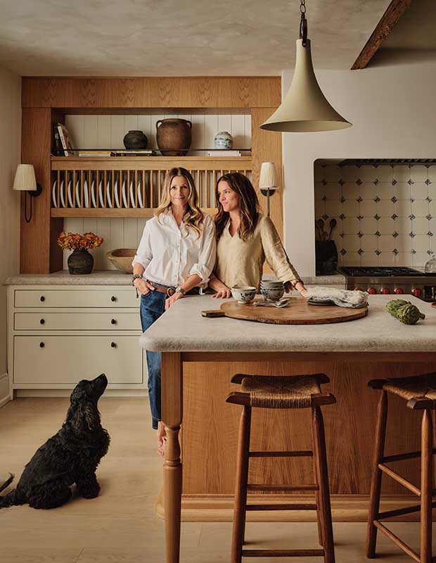 A photograph of two women standing at a kitchen island with a dish rack on the wall behind them and a black dog sitting on the ground looking up at them.
