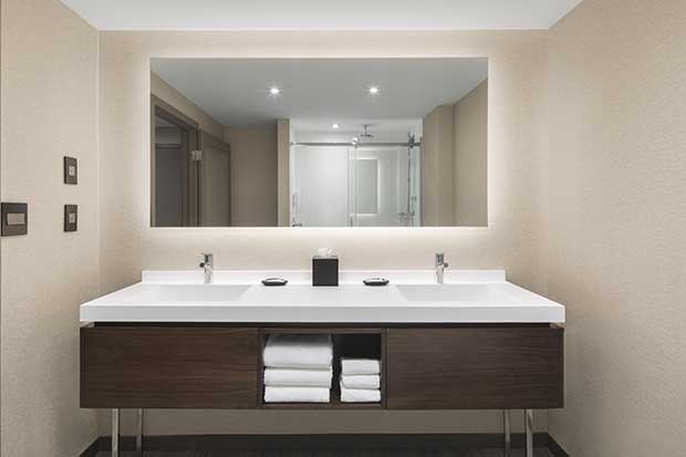 An image of a bathroom at the AC Old Montreal hotel. A vanity with brown wood cabinets and a white countertop sits below a mirror reflecting a shower on the opposite wall.