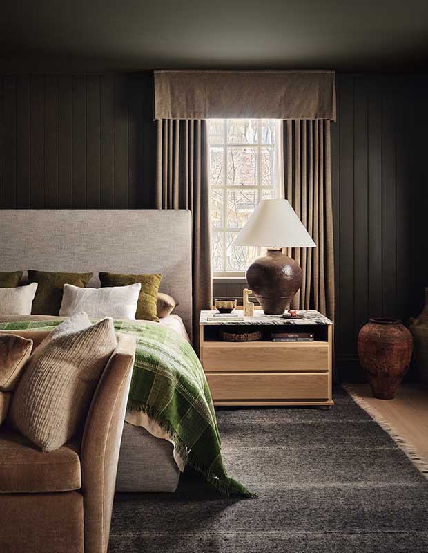 A photograph of the bedroom. On the left is the bed with a grey headboard and a green blanket covering it. Beside it is a bedside table with a round lamp on it in front of a window.