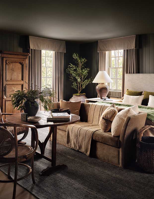 A photograph of the bedroom. On the rights, a bed sits against the wall, and a couch sits at the foot of the bed in front of a table with a book on it. The room is painted a dark, purple-y brown is illuminated by two windows.
