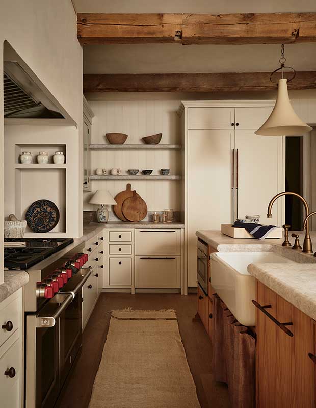 A photograph of the kitchen. White shelves and cabinets cover the back wall, while on the left, a range with red knobs sits under a white range hood, and on the right, a kitchen island features wood cabinets, white countertops and a farmhouse sink.