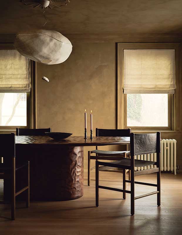 A photograph of a dining room with a large circular table made of hammered walnut with a reddish stain. A circular light fixture hangs overhead and the wall is textures with yellow limewash paint.