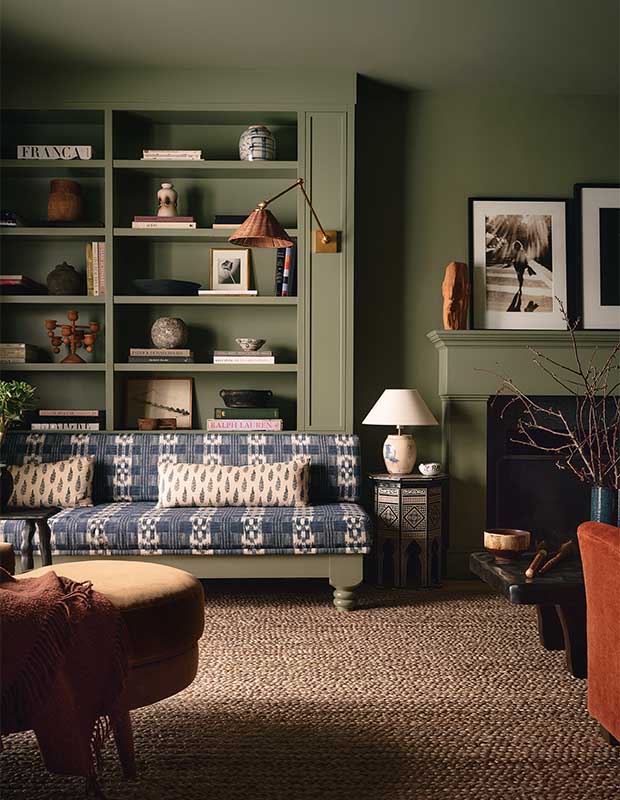 A photograph of the living room with green walls and a reddish carpet. A blue patterned couch sits with its back against a bookshelf with a sconce above it.
