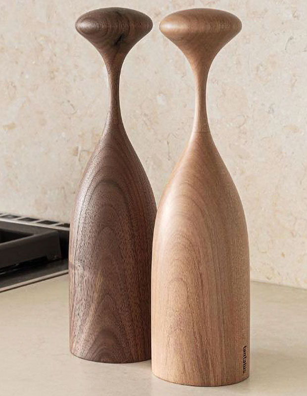 A photograph of a sustainable gift: two wood salt and pepper mills, one dark and one light, sit on a beige counter against a beige background. They are curved with very narrow necks.