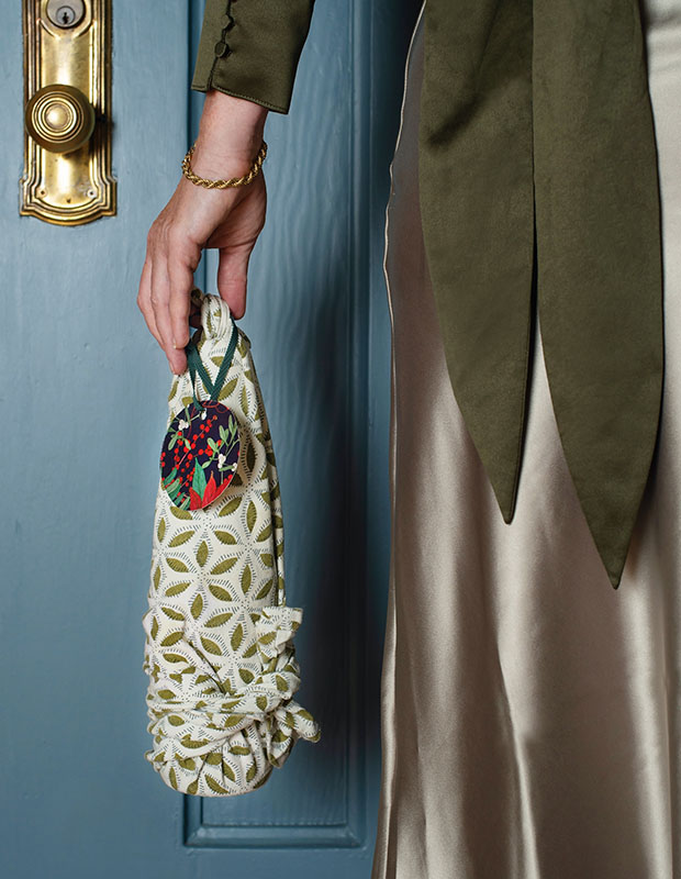 A photograph of a holiday craft: a bottle covered in a patterned dishcloth being held by someone wearing a green coat standing in front of a blue door.