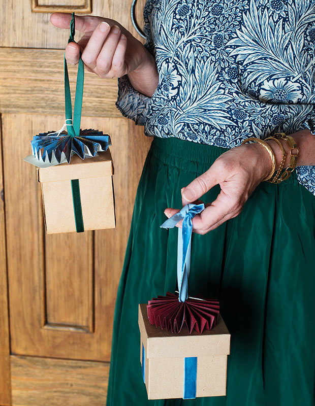 A photograph of a holiday craft: a person wearing a blue shirt and a green skirt is holding two ornaments in the shape of Christmas presents.