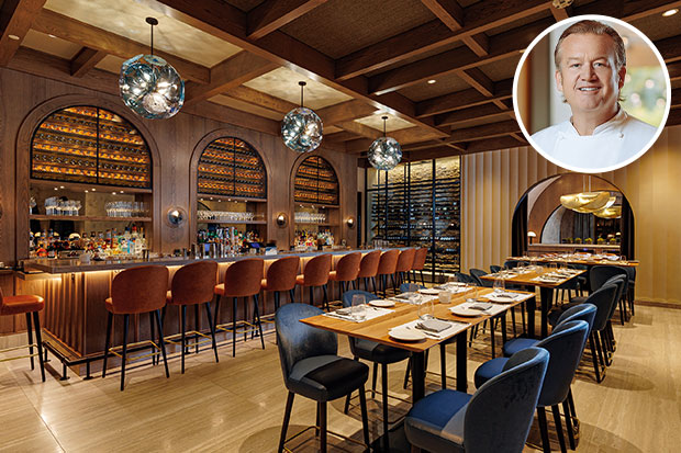Explore Nassau: A photograph of a dining room in a restaurant. In the foreground to the right, wooden tables are lined with blue chairs and set with white plates. In the background, a row of brown bar stools line a bar with disco balls hanging overhead. The walls and ceiling are warm wood. A photo of a middle-aged white man with blond hair (the chef) is inset in the top right corner.