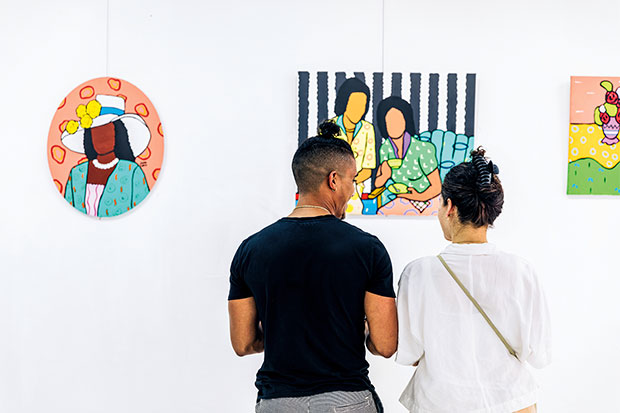 Explore Nassau: A photograph of two people with top buns face away from the camera in front of a piece of art hanging on a white wall. The person on the right is wearing a black t-shirt and is looking to the person beside them. The person on the right is wearing a thin white shirt and has a shoulder strap from a bag across their back. The piece of art features two human figures without facial features against a background of black and white.