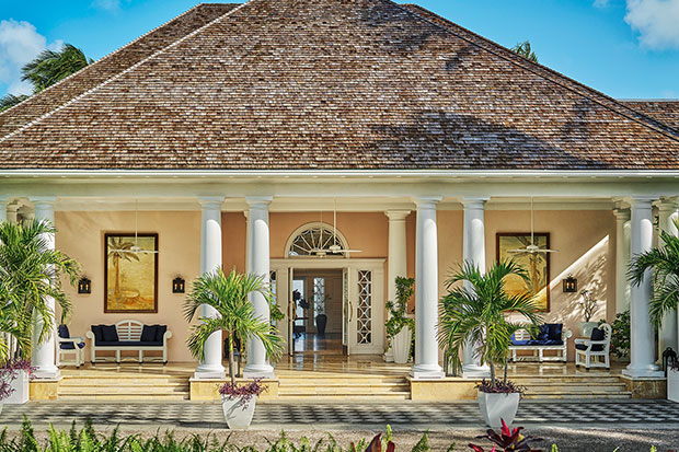 Explore Nassau: A photograph of a hotel entrance. The walls are yellow and the doorframe is white. A curved transom window sits above the door, and four white columns and two green, leafy potted plants sit on either side of it. A brown shingle roof rises above the veranda in a triangle against the blue sky.