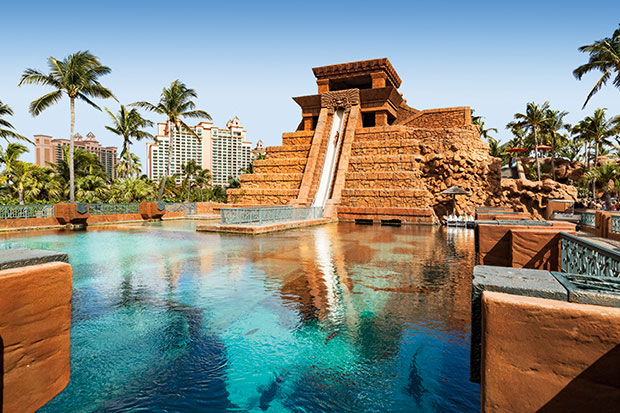 Explore Nassau: A photograph of a waterslide that is build into a reproduction Mayan temple. It runs down the side of the temple and then goes underneath a vivid-blue aquarium where the silhouettes of large fish are visible. In the background, palm trees and the top of a resort hotel is visible.