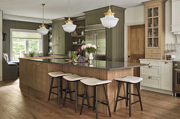 Kitchen with farmhouse aesthetic: A photo of a kitchen island with three pendants hanging above and four stools. Green cabinets and a fridge line the wall behind it. The island is wood with a shiny stone countertop.