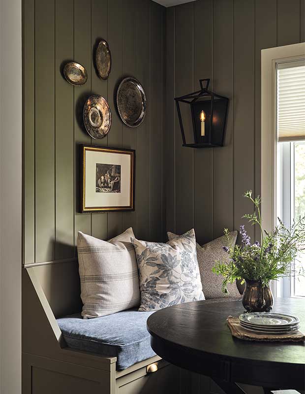 Kitchen with farmhouse aesthetic: A banquette is full of white pillows and a black table sits in front of it. The walls behind it are green and mounted with decorative plates, a picture frame, and a sconce.