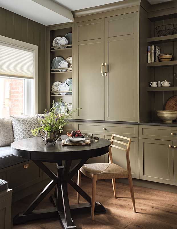 Kitchen with farmhouse aesthetic: A chair made of light wood sits at a dark, round table. Green cupboards sit behind it.