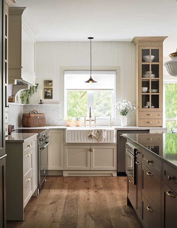 Kitchen with farmhouse aesthetic: A view of the farmhouse sink with a window above it and countertops on either side. In the foreground, a kitchen island is on the right and cupboards are on the left.
