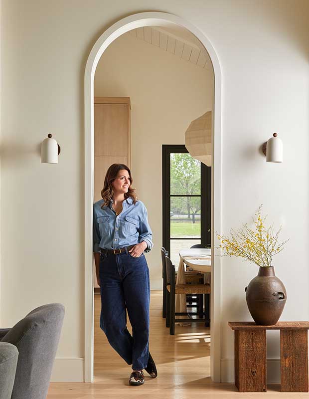 Photo of house featuring English design: A woman in a blue shirt and blue jeans with blond hair stands in an arched doorway. A vase sits beside her.