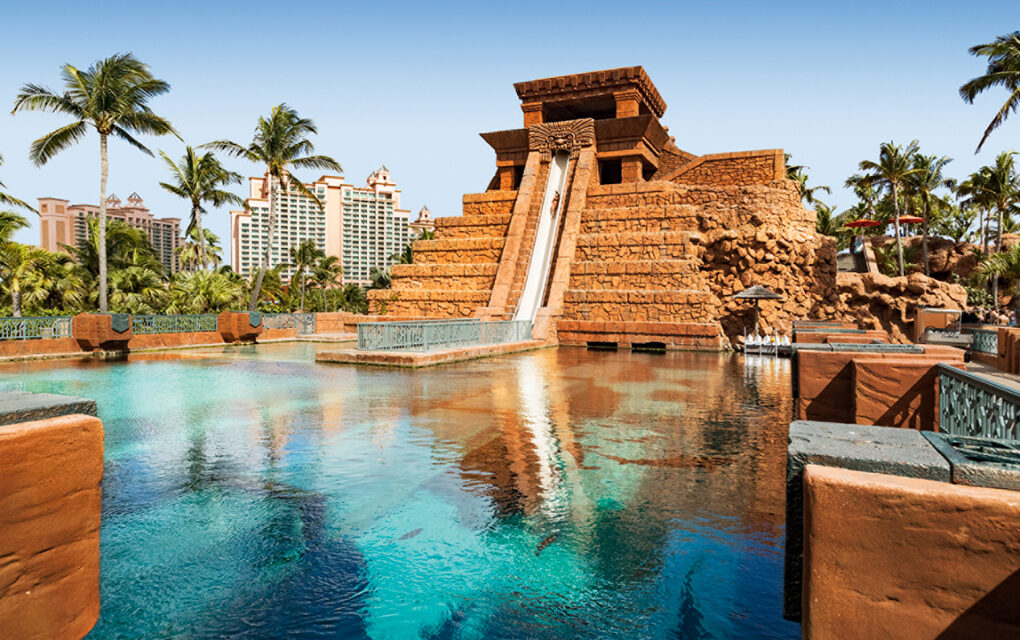 Explore Nassau: A photograph of a waterslide that is build into a reproduction Mayan temple. It runs down the side of the temple and then goes underneath a vivid-blue aquarium where the silhouettes of large fish are visible. In the background, palm trees and the top of a resort hotel is visible.