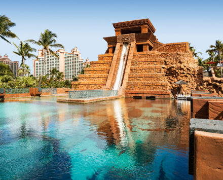 Explore Nassau: A photograph of a waterslide that is build into a reproduction Mayan temple. It runs down the side of the temple and then goes underneath a vivid-blue aquarium where the silhouettes of large fish are visible. In the background, palm trees and the top of a resort hotel is visible.